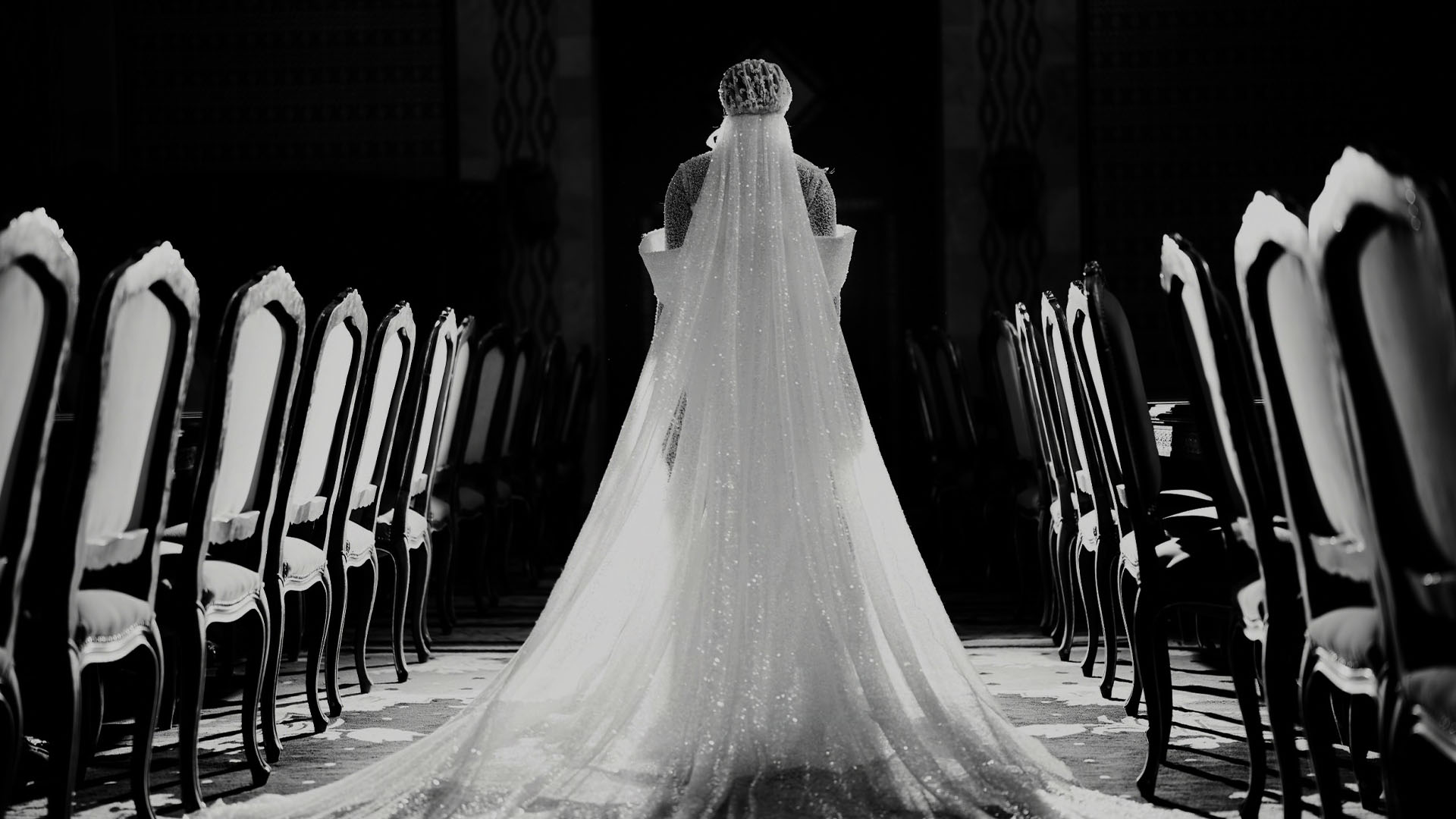 Back view of a bride in a long, sparkling wedding gown standing between rows of chairs in a dark room.