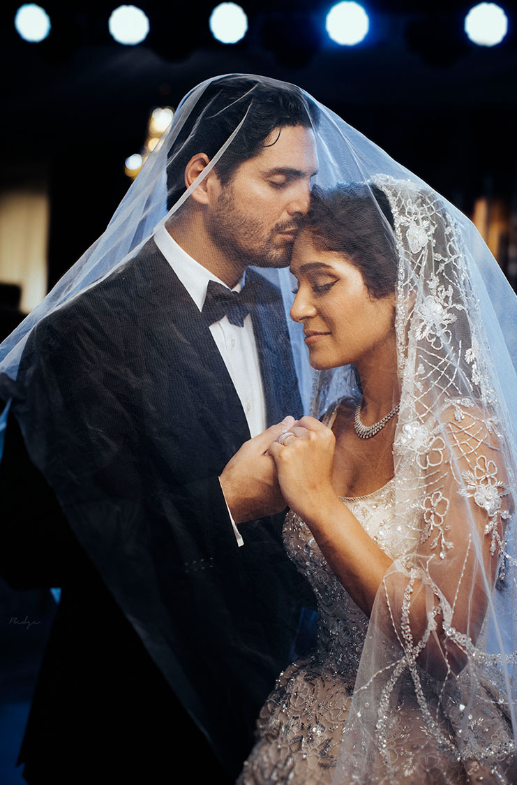 Bride and groom embracing under a sheer lace veil, holding hands with eyes closed in a tender moment.