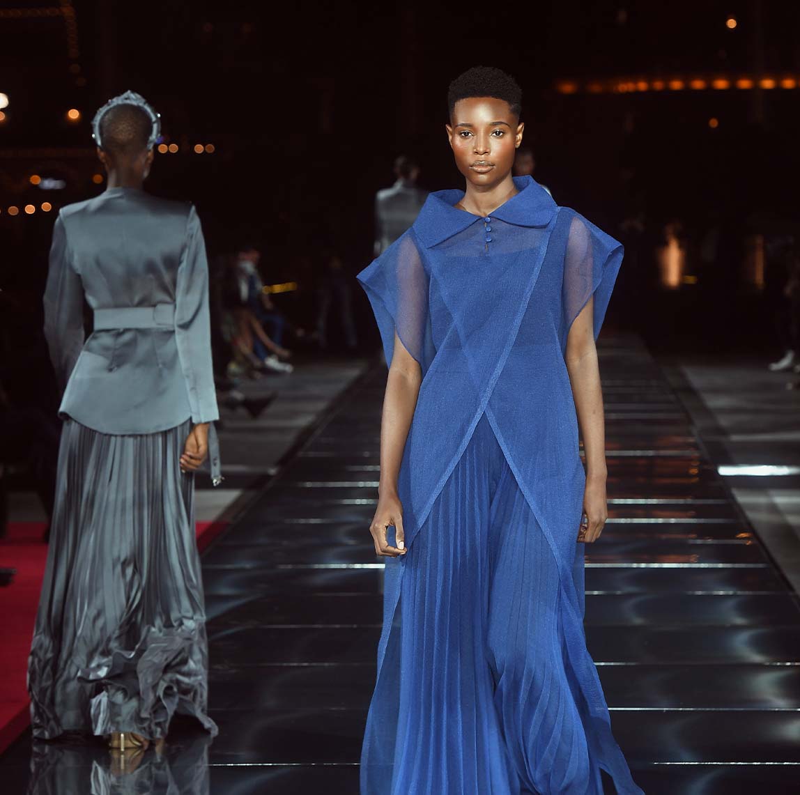 Model in a sheer blue dress walking on a runway during a nighttime fashion show.