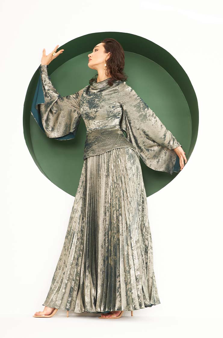 Woman in a long, flowing silver-grey pleated dress with wide sleeves posed against a green circular backdrop.