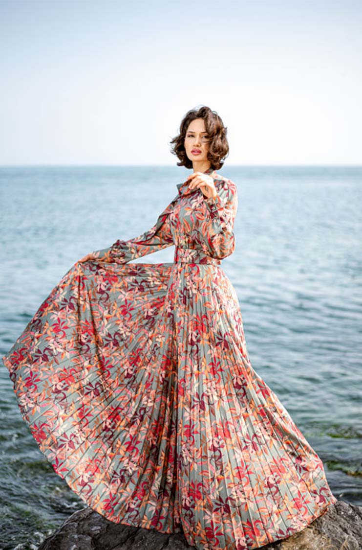 Woman in a long, floral-patterned dress standing on rocks by the sea, holding the dress out to display its flow.