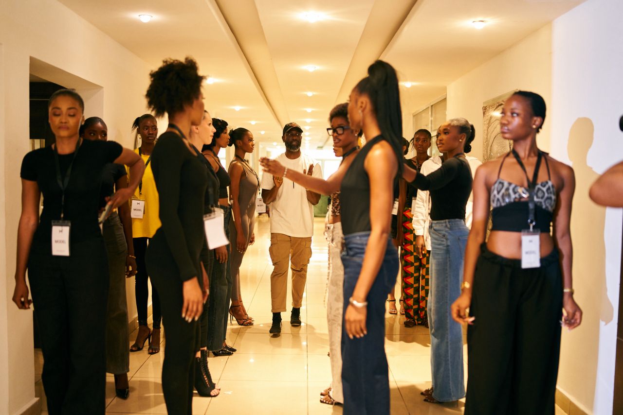 Fashion models lined up facing each other in a hallway with a man walking between them.